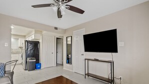 Sala de estar con televisor, ventilador de techo, una mesa de madera y una zona de cocina al fondo.