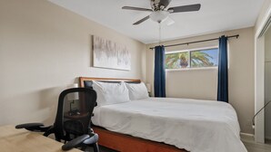 Un dormitorio con una cama, un escritorio, una silla, un ventilador de techo y una ventana con cortinas.