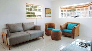 A living room with a grey sofa, two wicker chairs with blue cushions, a glass coffee table, and a large window with a view of a balcony. - Casa del Sol (Noord)
