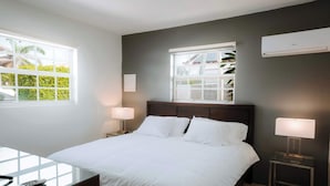 A hotel room with a large bed, two bedside tables, a nightstand, a window with a view of greenery, and an air conditioning unit. - Casa del Sol (Noord)