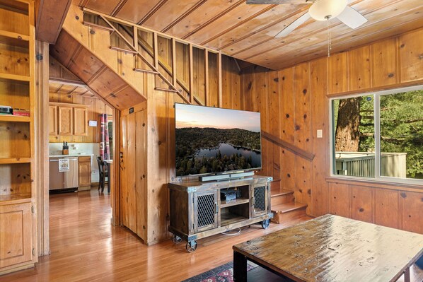 A wooden interior with a TV on a cart, a staircase, and a view of a forest through a window.