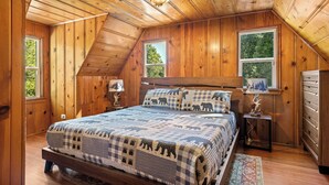 A bedroom with a wooden ceiling, a bed with a checkered quilt, two windows, and a nightstand with a lamp.