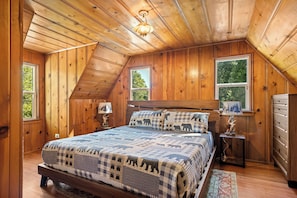 A bedroom with a wooden ceiling, a bed with a checkered quilt, two windows, and a nightstand with a lamp.