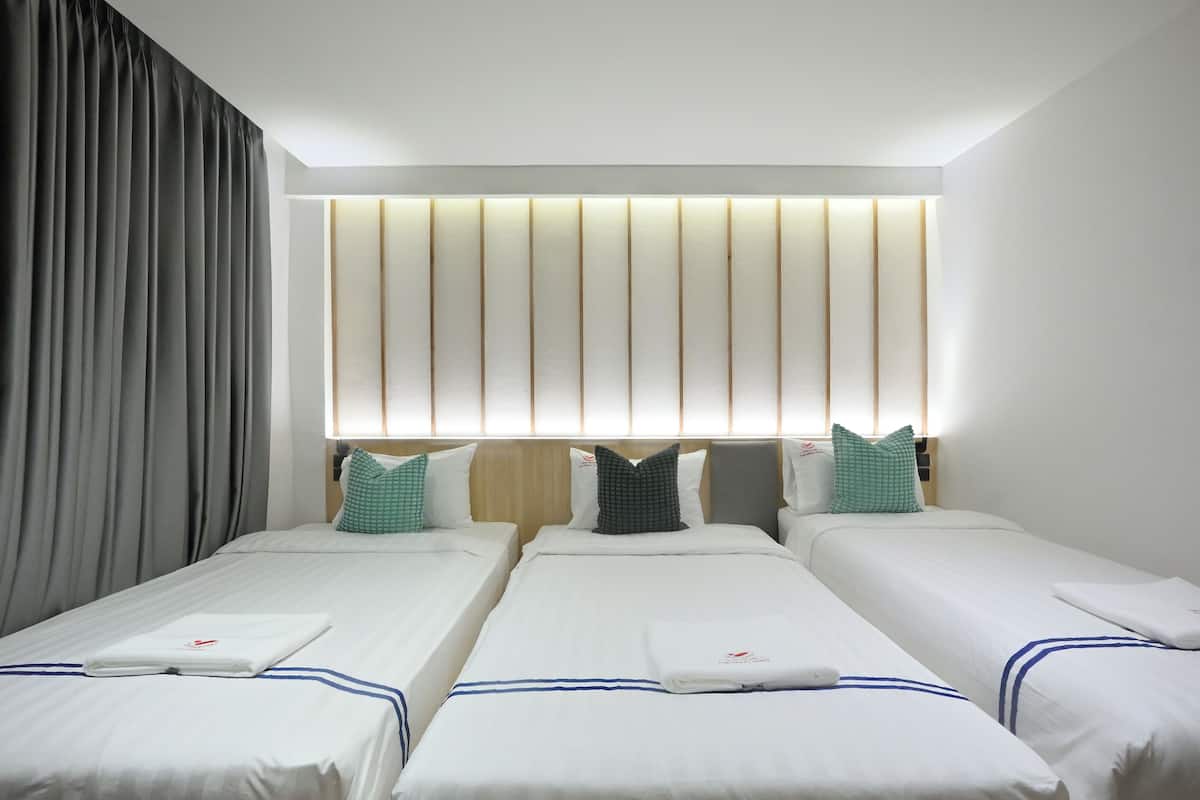 A hotel room with three single beds, a large window with vertical blinds, and a modern headboard with integrated lighting.