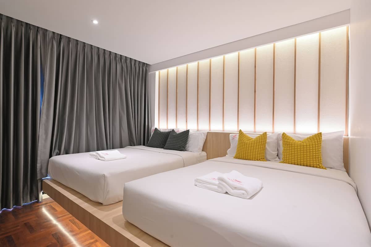 A hotel room with two beds, a wooden floor, a large window with curtains, and a wall-mounted light panel.