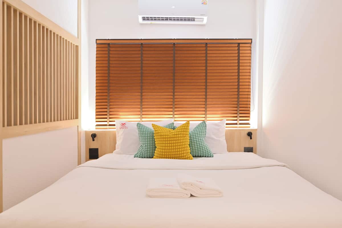 A neatly made bed with a white duvet, a set of folded towels, and a window with wooden blinds.