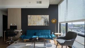 Een moderne woonkamer met een blauwe bank, een salontafel, een bijzettafeltje en een vloerlamp. Er is een groot raam met zonwering en een stadsgezicht is zichtbaar buiten.