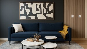 Un sofà blau fosc amb un coixí, dues taules rodones amb sobre de marbre, un ramader amb flors seques i un quadre abstracte emmarcat a la paret.
