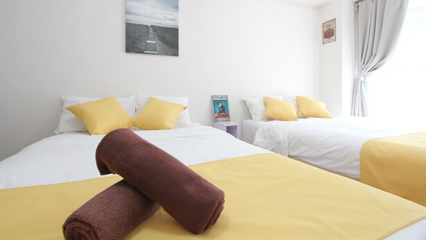 Una habitación de hotel con dos camas, cada una con ropa blanca y almohadas amarillas. Hay una toalla marrón enrollada sobre la cama en primer plano.