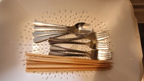 Un colador blanco con varios utensilios, como cucharas, tenedores y palillos, dispuestos sobre él.