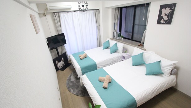 A hotel room with two beds, a TV, a clock, a window with curtains, and a plant.