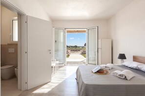 A bedroom with a bed, a nightstand, a lamp, a view through an open door to an outdoor patio, and a bathroom with a toilet and sink.