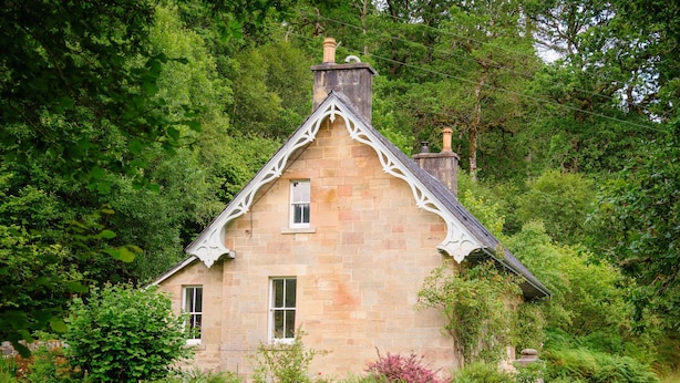 A stone house with a gabled roof, surrounded by lush greenery and a stone wall.