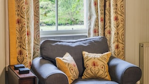 A room with a grey sofa, two patterned cushions, a small wooden table with a stack of books, and a window with floral curtains.