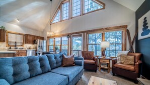 Ein geräumiges Wohnzimmer mit einer großen blauen Couch, einem hölzernen Couchtisch und einem braunen Ledersessel. Mehrere Fenster sorgen für natürliches Licht und Ausblicke ins Freie.