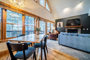 A spacious living room with a large wooden dining table, blue upholstered chairs, a grey sofa, and a stone fireplace.
