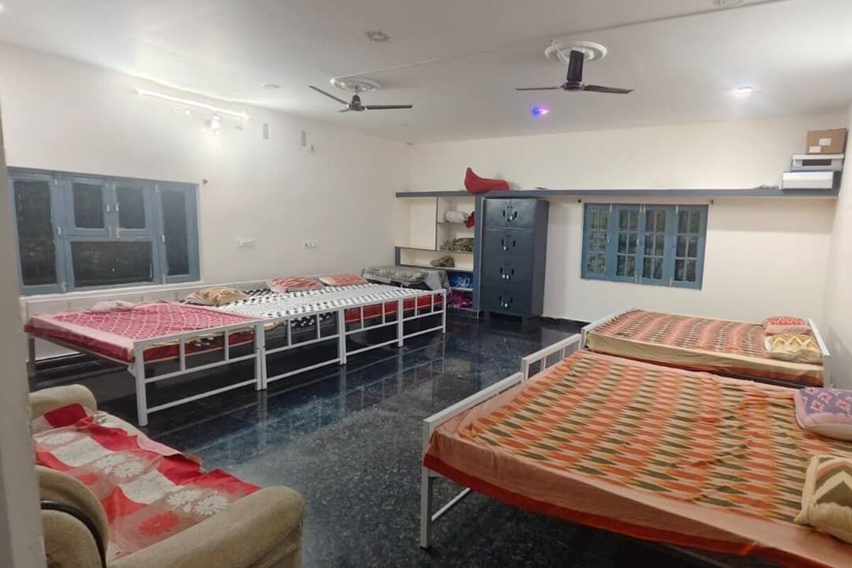 A dormitory room with three beds, a wardrobe, a desk, and a ceiling fan.