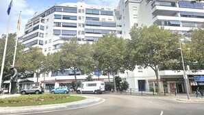 Un edifici de diverses plantes amb balcons, una estàtua i un carrer amb cotxes i vianants.
