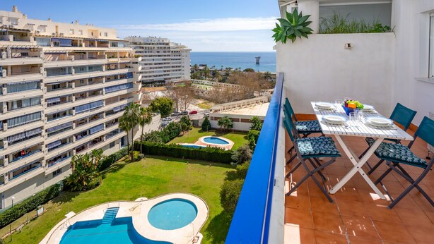 Un balcó amb taula i cadires, una piscina i vista al mar i edificis.