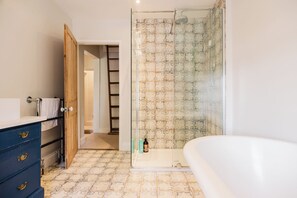 A bathroom with a bathtub, a glass shower enclosure, a wooden door, and a tiled floor. - Historic 3 BD Cottage in the Cotswolds (Milton-under-Wychwood)