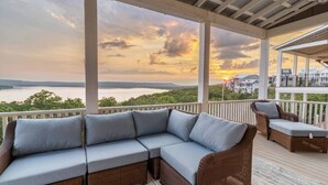 A covered outdoor seating area with a wicker sofa and chairs, overlooking a body of water and distant buildings. - Shore Enough | Carlton Landing Lakeview, Up to 18 (Eufaula)