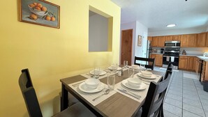 A dining area with a wooden table set for four, a painting of oranges on the wall, and a kitchen in the background. - 3Br Farmington Home & 75” Smart Tv + Pet Friendly (Farmington)