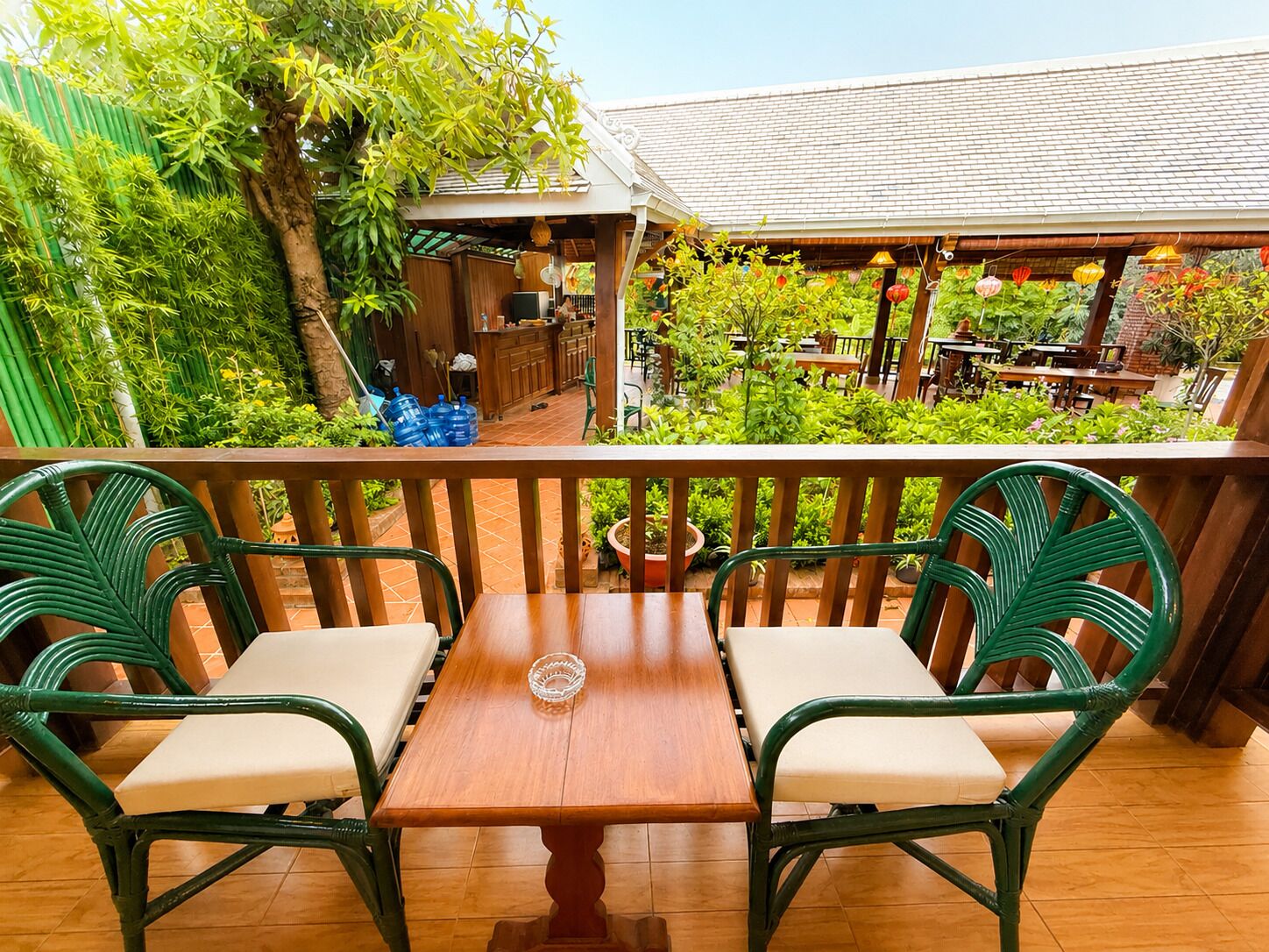 A balcony with two green wicker chairs and a wooden table, overlooking a garden with various plants and a wooden structure in the background.