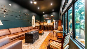 A cozy living room with a leather sofa, a coffee table, and a large window overlooking a deck. - Newly Renovated, Cozy Mid-Lake Cabin; Sleeps 10 (Many)