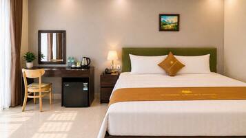 A hotel room with a bed, a desk, a chair, a small refrigerator, a lamp, and a painting on the wall.