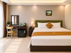 Ein Hotelzimmer mit Bett, Schreibtisch, Stuhl, kleinem Kühlschrank, Lampe und einem Gemälde an der Wand.