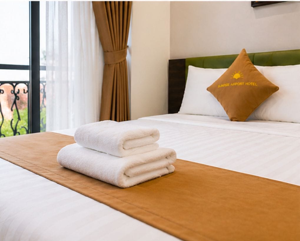A neatly made bed with white linens and a brown headboard, a pillow with a logo, and a window with curtains.
