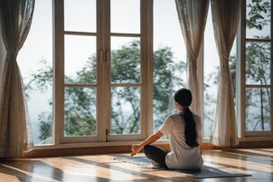 Eine Person sitzt auf einer Yogamatte in einem Raum mit großen Fenstern und Vorhängen.
