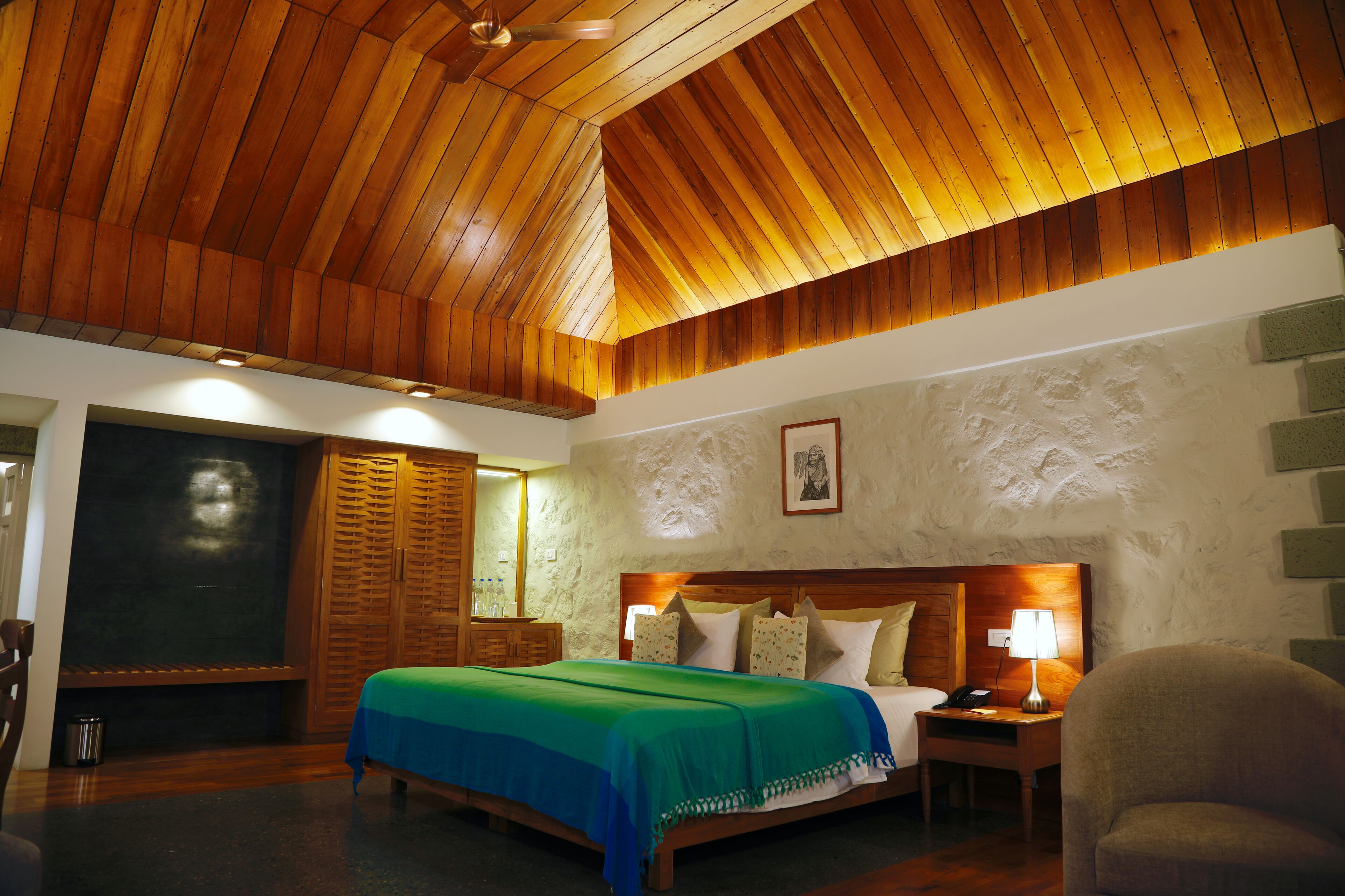 Ein Schlafzimmer mit Holzbalkendecke, einem Bett mit grüner Decke, einem Nachttisch mit Lampe und einem Sessel.