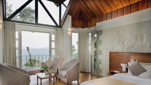 A bedroom with a large bed, two armchairs, a small table, and a view of the outdoors. - SpiceTree Rajakumari (Udumbanchola)