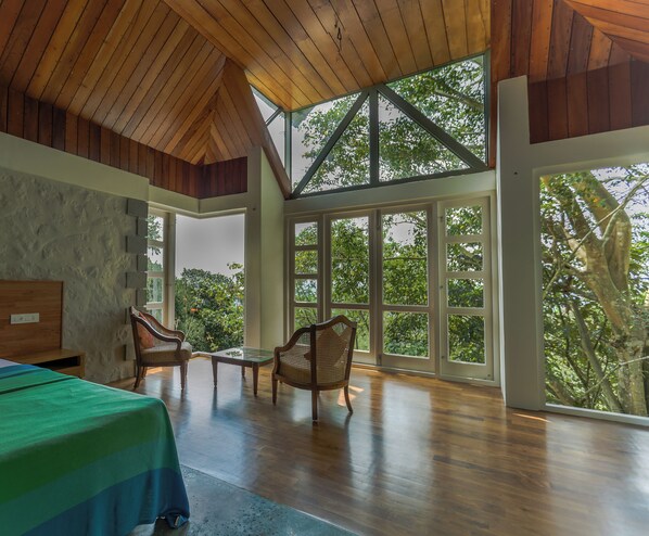 A spacious room with a high ceiling and large windows offering a view of trees. There are two wooden chairs and a table.