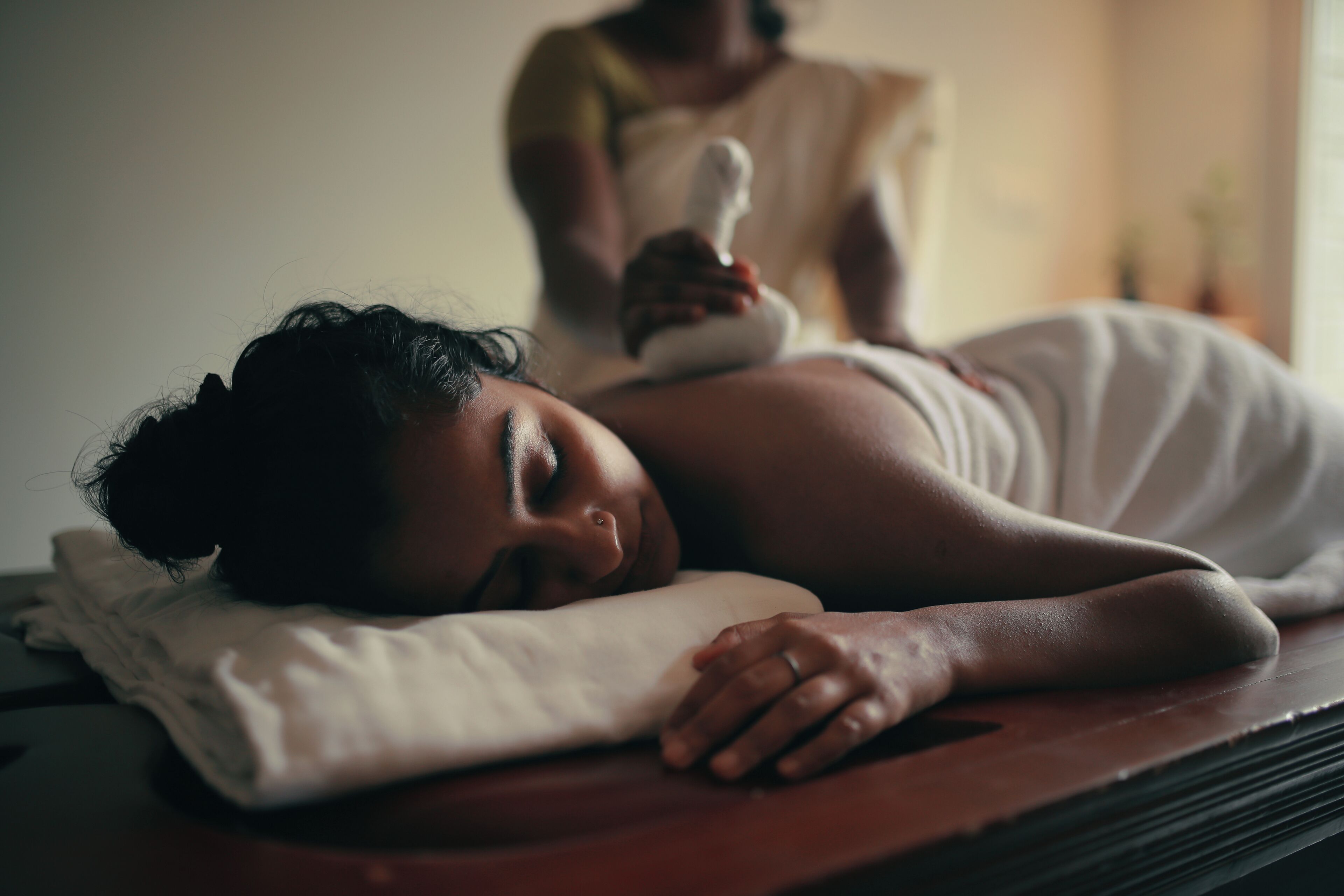 A person lying on a massage table with a towel covering their body, in a room with a wooden floor and a plain wall in the background.