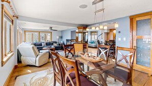 A spacious living room with a glass dining table, wooden chairs, a sofa, and a kitchen area in the background.