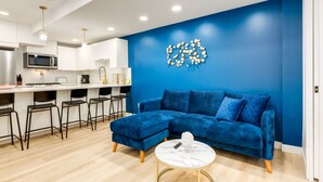 Uma sala de estar moderna com um sofá azul, uma mesa de centro branca e uma área de cozinha com armários brancos e um micro-ondas.