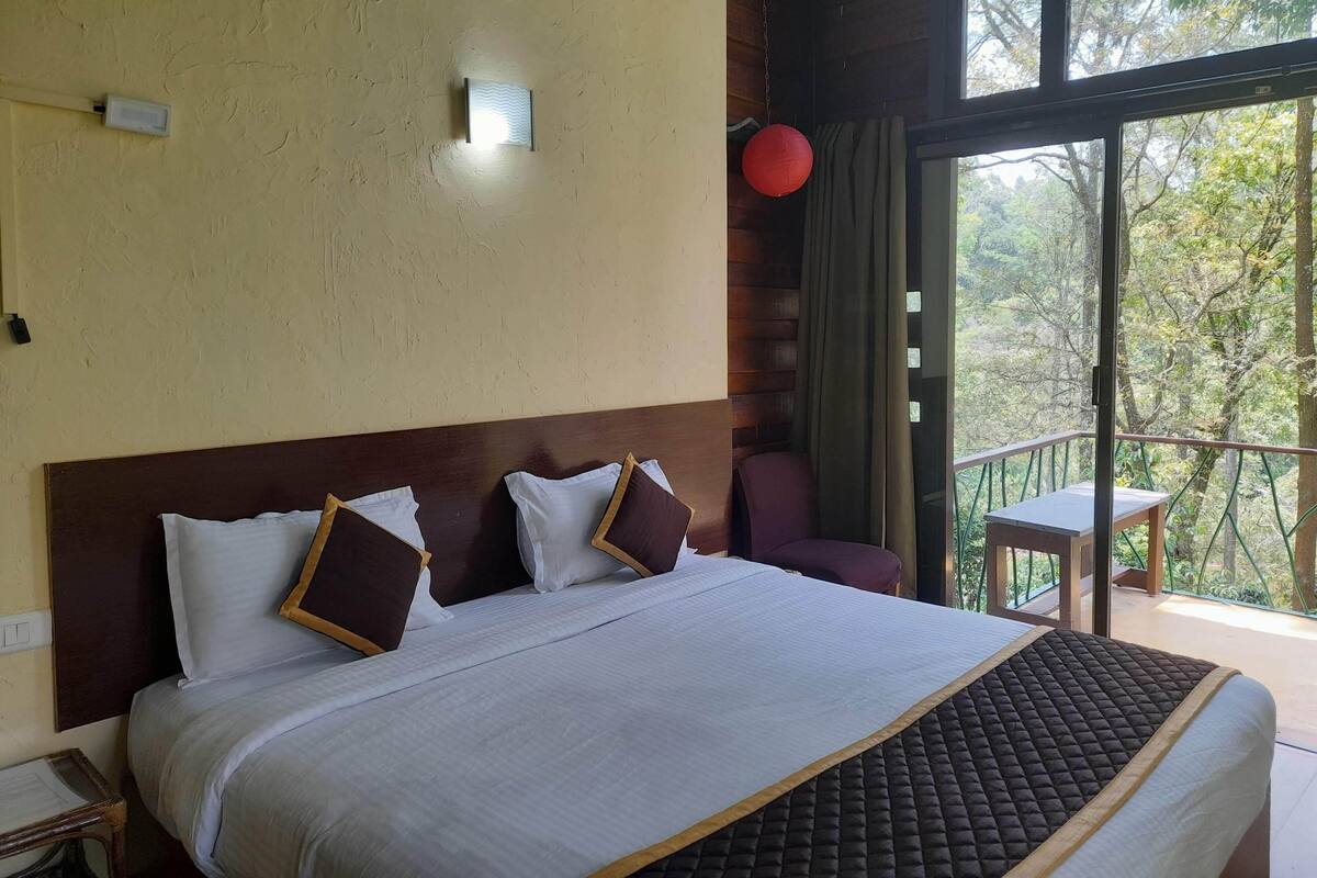 Habitación de hotel con una cama grande, cabecera de madera, mesita de noche, un farol rojo y un balcón con vistas a árboles.