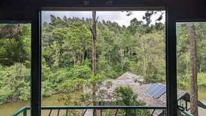 A view from inside a building looking out at a forested area with a water body and a small structure with solar panels. - Gorukana Eco Wellness Centre (Yelandur)