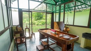 A traditional wooden table with a hanging pot, a small stool, and a chair in a room with large windows and a green roof. - Gorukana Eco Wellness Centre (Yelandur)