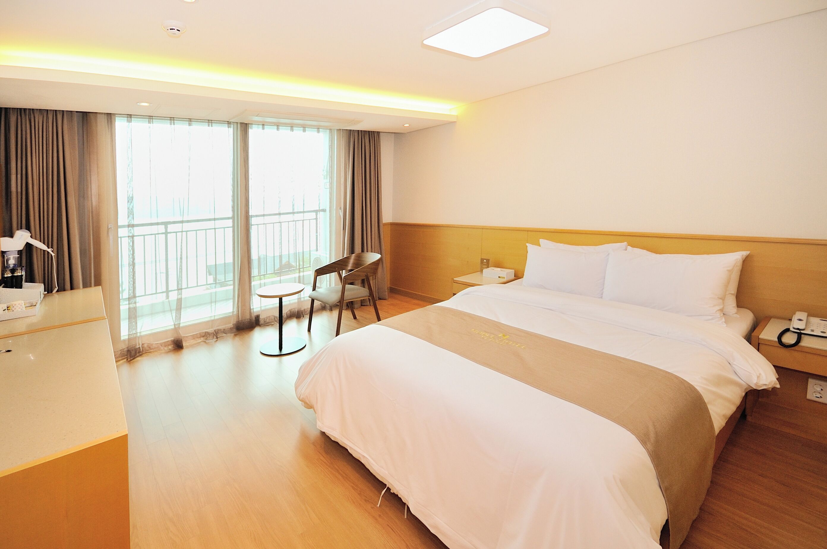 A hotel room with a large bed, a desk, a chair, and a balcony with curtains.