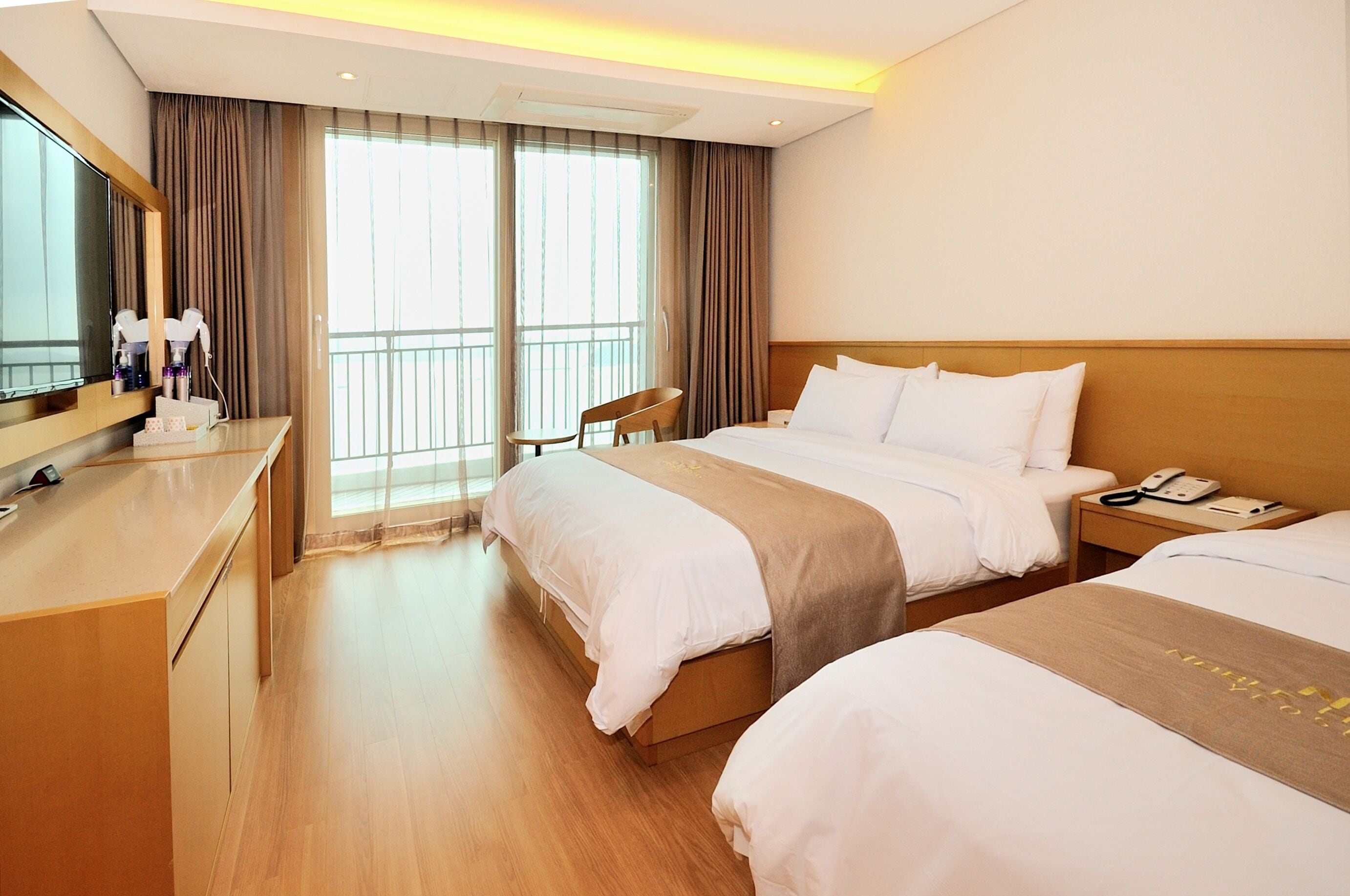 Habitación de hotel con dos camas, un escritorio, una silla, televisión y balcón con cortinas.