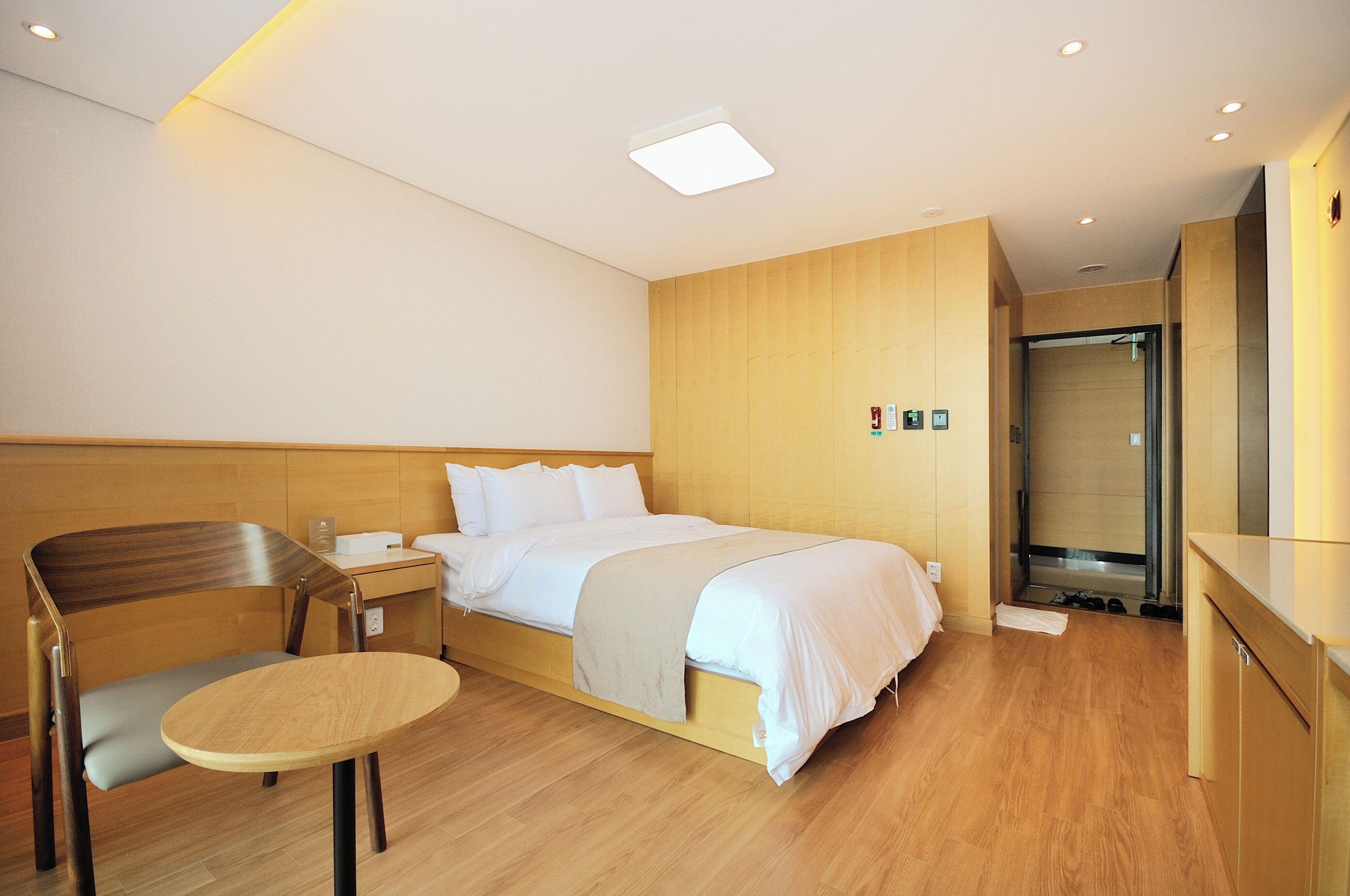 Una habitación de hotel moderna con una cama de madera, una mesita redonda pequeña, una silla y un espacio con ducha.