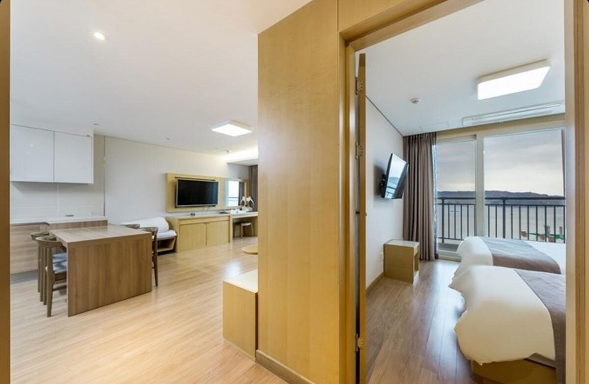 A modern hotel room with a wooden floor, a balcony with a view, a kitchen area, and a TV.