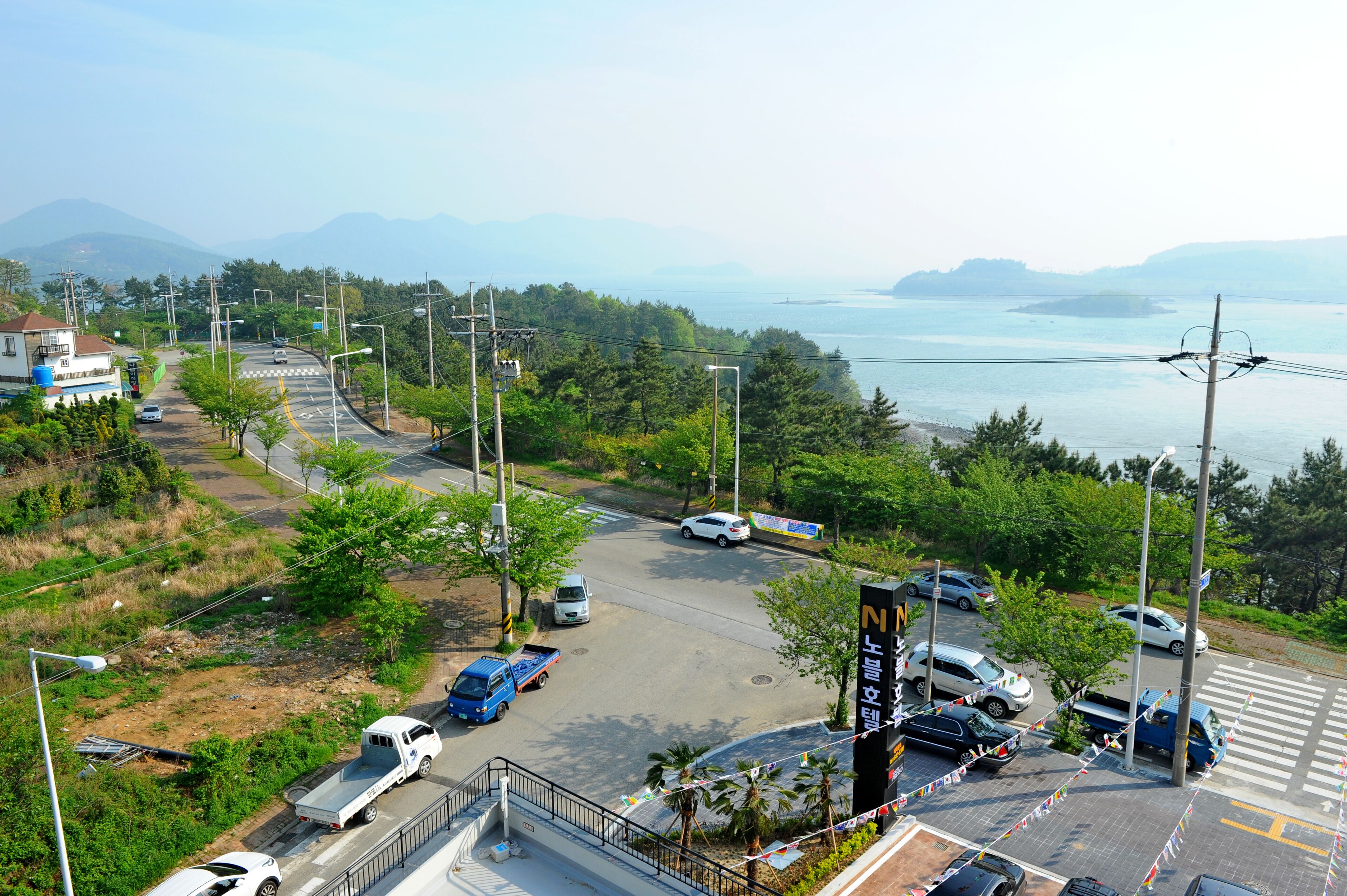 A parking lot with multiple cars and a blue truck, surrounded by greenery and a view of a body of water and mountains.