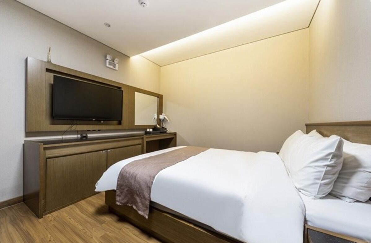 A hotel room with a bed, a flat-screen TV mounted on a wooden cabinet, a bedside table with a lamp, and a large mirror above the TV.