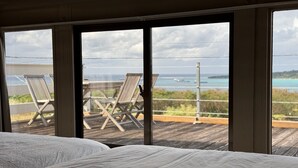 A bedroom with a large window overlooking a deck with two chairs and a table. - Private Villa with Ocean Views | Churaumi & JUNGLIA | BBQ & Fireworks | Sleeps 9 (本部町)