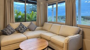 A modern living room with a beige sectional sofa, two patterned throw pillows, a wooden coffee table, and large windows offering a view of greenery and the sky. - Private Villa with Ocean Views | Churaumi & JUNGLIA | BBQ & Fireworks | Sleeps 9 (本部町)