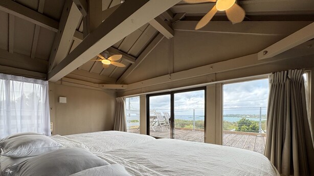 A bedroom with a large bed, a ceiling fan, and a view of the sea through large windows. - Private Villa with Ocean Views | Churaumi & JUNGLIA | BBQ & Fireworks | Sleeps 9 (本部町)